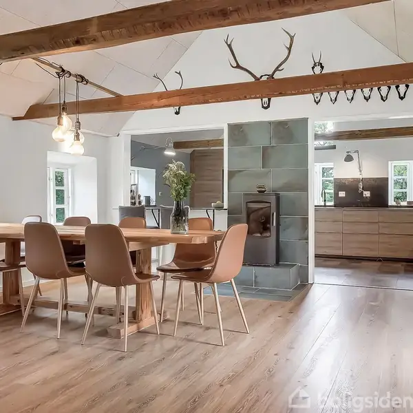 Et stort træbord med otte stole står i en lys spisestue. Over bordet hænger en lampe, og væggen bagved har en ovn med fliser. Køkkenet ses i baggrunden.