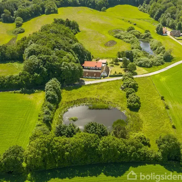 En stor gård med rødt tag ligger omgivet af frodige grønne marker og træer. En lille sø ses foran gården, og en vej fører til ejendommen gennem det landlige landskab.