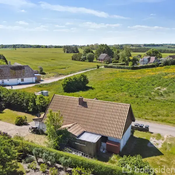 En gårdbygning med rødt tag står stille i et landligt område omgivet af grønne marker og få træer under en klar blå himmel. Tre mindre bygninger ses i baggrunden.