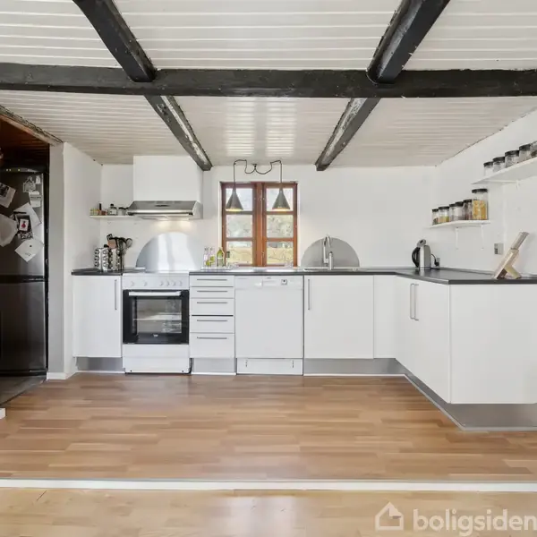 Et køkken med hvide skabe og sort bordplade. Træbjælker i loftet. Køleskab, ovn og vask er synlige. Der er krydderiglas på hylderne og trægulve. En lille vindue centralt bagtil.