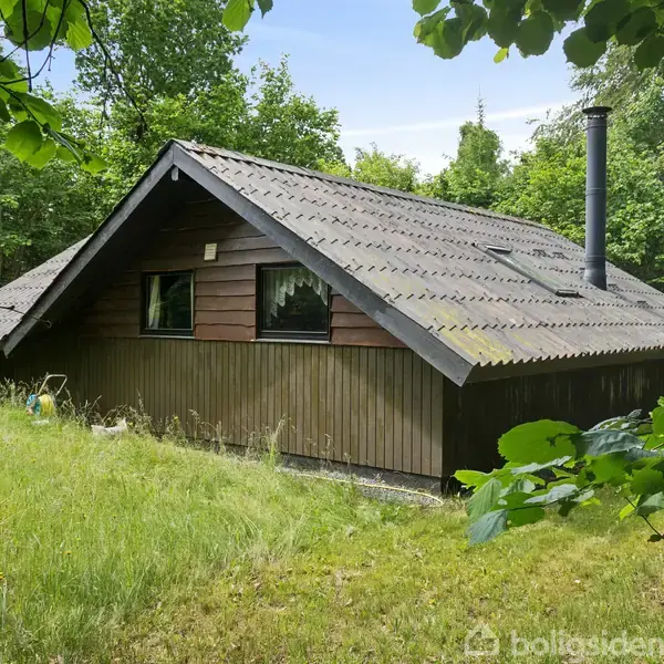 Et lille træhus står stille i en frodig have omgivet af tætte træer. Huset har skrånende tag med en skorsten. Sollys skinner gennem de grønne blade.
