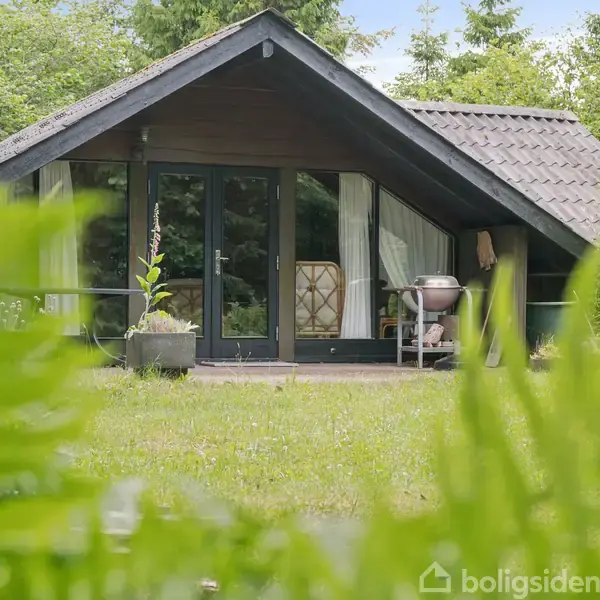Sommerhus med store glasdøre og en grill på terrassen, omgivet af frodig grøn bevoksning.