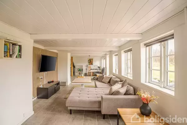 Stor grå sofa står i en lys og rummelig stue med hvidmalede trælofter og flisegulv. Væggene har store vinduer, og et vægmonteret TV hænger til venstre.