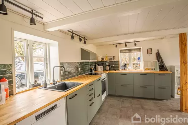 Køkken med grå skabe og træbordplader, udstyret med komfur og vask. Rummet er lyst med vinduer, og der er køkkenudstyr på bordpladen. Fliser i grøn nuancer pryder væggen.