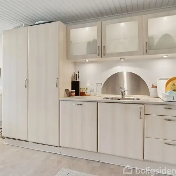 Køkken i lyse nuancer med skabe og skuffer, opvaskemaskine under vasken samt tallerkener ved siden af. Omgivet af hylder med forskellige køkkenredskaber.