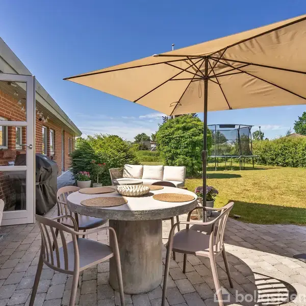 Terrasse med et rundt betongbord omringet af stole under en stor parasol; i baggrunden ses en grøn have med en trampolin.