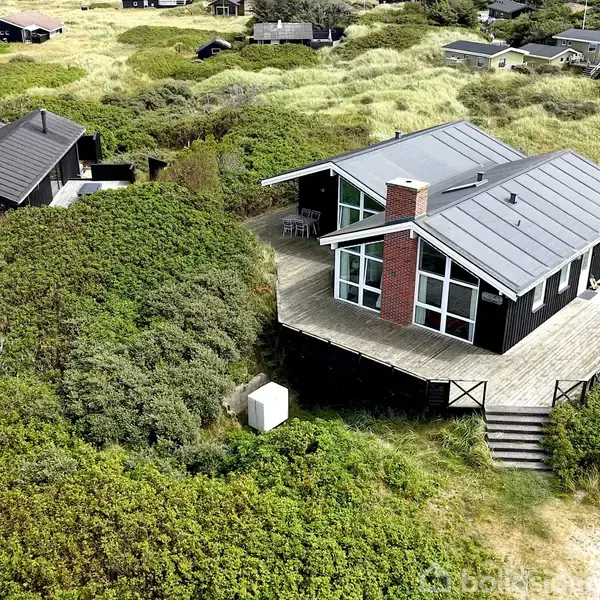 Sort sommerhus stående stille på stor terrasse, omgivet af frodige klitter og grøn vegetation. Flere lignende huse kan ses i afstanden, indlejret i det naturskønne klitlandskab.