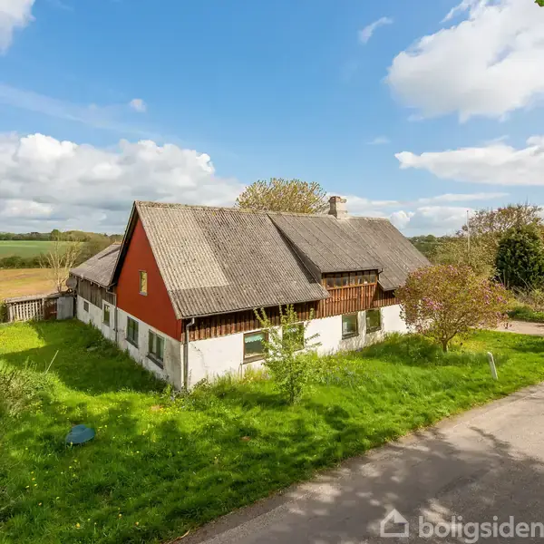 Gammelt stråtækt hus står på en grøn, frodig grund omgivet af en vej og marker under en blå himmel med spredte skyer.