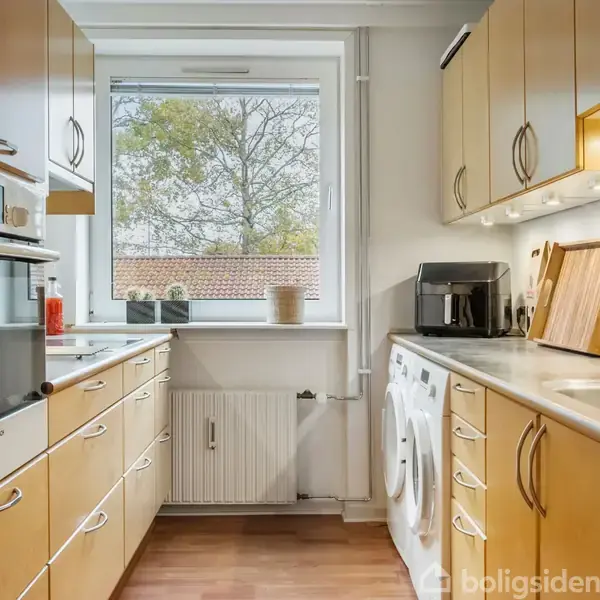 Et lyst køkken med træskabe og integrerede hvidevarer, herunder en ovn og vaskemaskine. Køkkenet har et vindue med udsigt til træer, og der ligger skærebrætter ved siden af vasken.