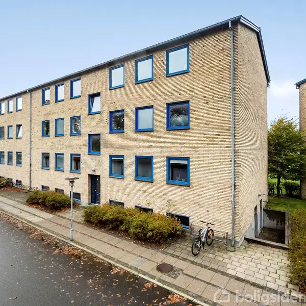 Et treetagers murstensbyggeri med blå vinduesrammer står langs en gade. En cykel er parkeret på fortovet, omgivet af buske. Det regnvåde fortov følger bygningens facade.