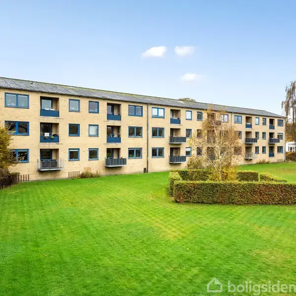 En treetagers gul murstensbygning med altaner står i en grøn græsplæne med velplejede hække. Bygningen har flere vinduer og er omgivet af et åbent, grønt område.