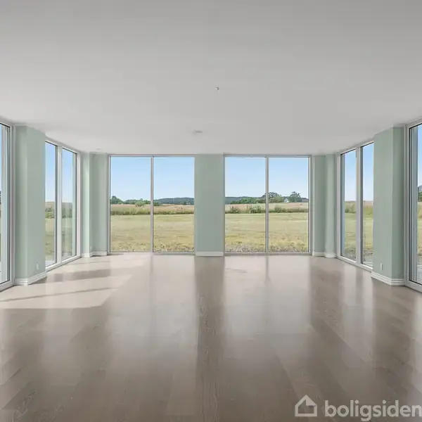 Et stort, tomt rum med trægulve; panorama-vinduer omkranser, giver udsigt til åbne, grønne marker under en klar blå himmel. Ingen tekst i billedet.