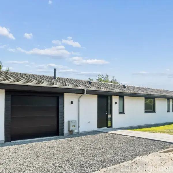 En moderne, hvidstens villa med sort tag står i en solrig gård. En grusbelagt indkørsel fører til en garage. Huset er omgivet af grønne områder under en klar himmel.