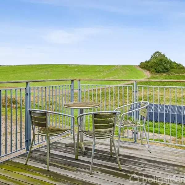 Tre metalstole og et bord står på en træterrasse. De er placeret med udsigt over et grønt landskab med en blå himmel i baggrunden.
