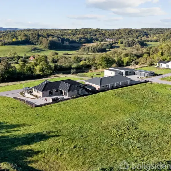 Huse ligger spredt på et grønt, bakket landskab. Omkringliggende skove og en klar himmel skaber en fredfyldt atmosfære. Husene er moderne og har veje imellem dem.