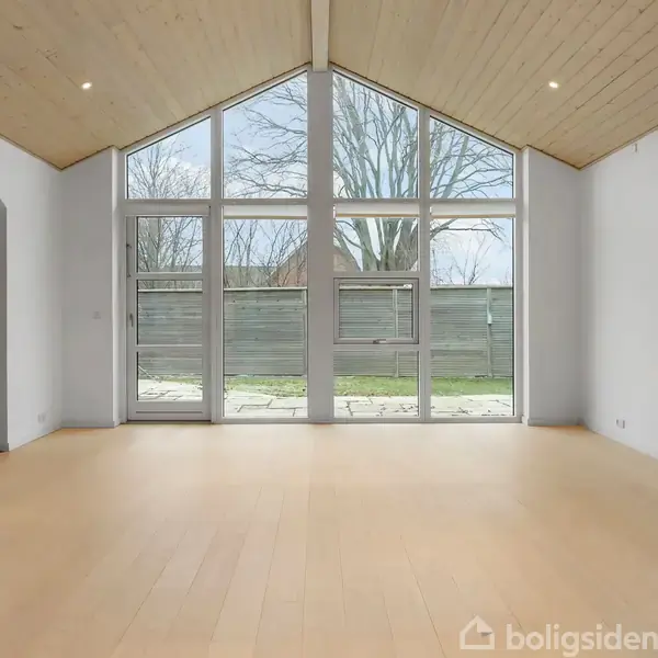 Et lyst og rummeligt rum med højt til loftet og store vinduer, der åbner op til en have. Trægulvet og det træbeklædte loft skaber en hyggelig atmosfære.