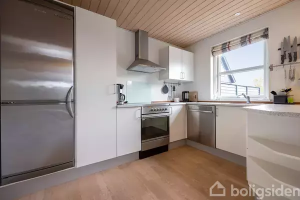 Et moderne køkken med hvide skabe indeholder et køleskab, ovn og emhætte. Rummet er lyst op af et vindue med udsigt, og gulvet er i lyst træ.