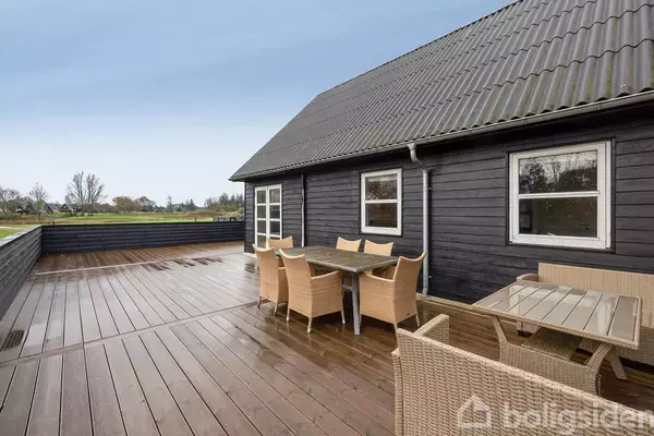 Træhus med mørke vægge, hvor der står havemøbler på en stor træterrasse. Terrassen er omkranset af et lavt hegn, og landskab ses i baggrunden.