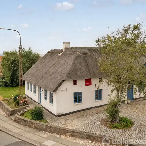 Hvidt bindingsværkshus med stråtag står i et hjørne af en brolagt gade omgivet af træer og huse. Huset har blå og røde detaljer og er omkranset af buske.