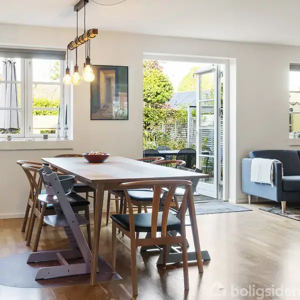 Spisebord med seks stole i en lys stue, hvor et åbent vindue afslører en have. Sofa og planter i baggrunden skaber en hyggelig atmosfære.