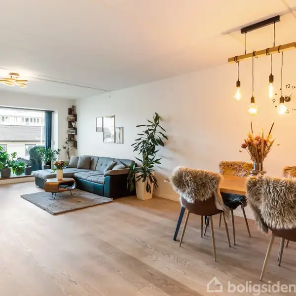 Stue med en lædersofa ved siden af planter og et sofabord på et tæppe. Et spisebord med stole dækket af pelspuder står under hængende lamper. På væggen står "We do what we love".