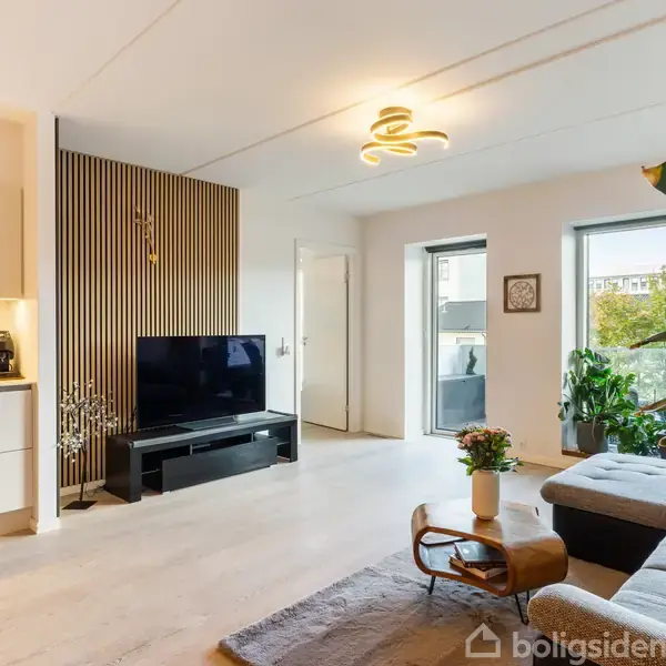 TV står på et lavt bord. I baggrunden ses en moderne stue med lyse vægge, store vinduer og planter. Til venstre er der et åbent køkkenområde.