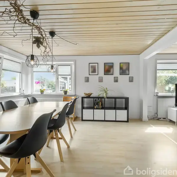 Spisebord med sorte stole står under en dekorativ lampe. Rummet har lyst trægulv og træloft, med billeder på væggen. Store vinduer giver udsigt og naturligt lys.