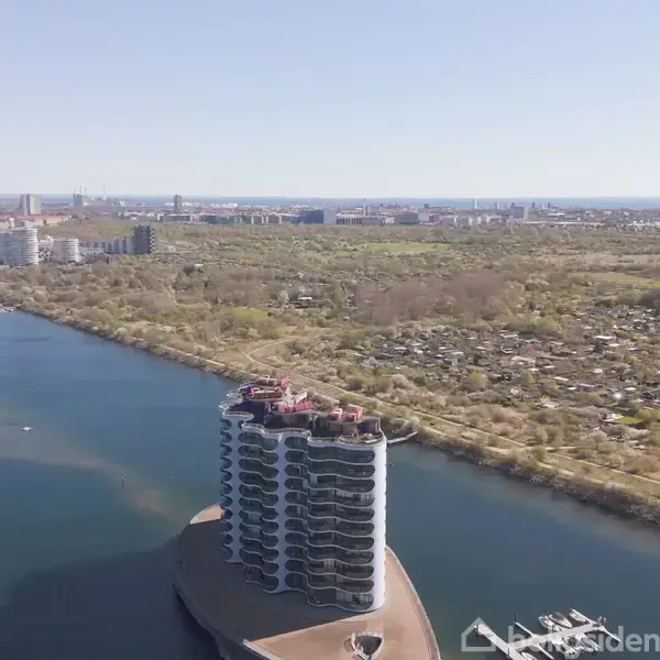 En høj bygning med balkoner strækker sig op mod himlen ved siden af en bred flod. Bygningen står på en halvø, omgivet af frodigt grønt landskab.