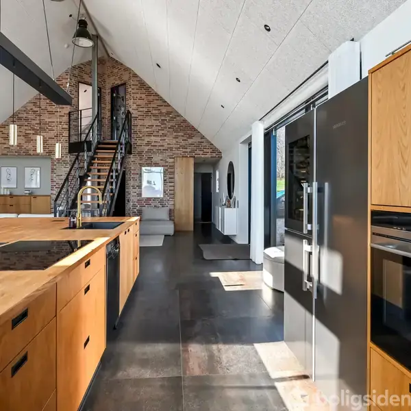 Træbordplade fungerer som central køkkenø, flankeret af moderne apparater. Trappe fører op til hemsen, mens vinduerne lader sollys strømme ind i det rummelige, moderne interiør med loft til kip.