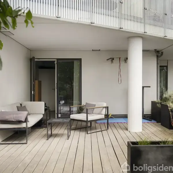Terrassen har moderne udemøbler, der står omkring et lille bord. Den er omgivet af grønne planter og ligger under en buet balkon, hvilket skaber en hyggelig atmosfære.