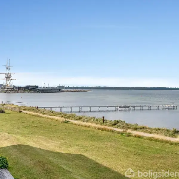 Et skib med høje master ligger til kaj ved en rolig kystlinje. En lang bro strækker sig ud i vandet, omkranset af grønt græs og lave buske.