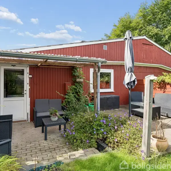 Et rødt hus med en terrasse af fliser foran, hvor der står udemøbler og en parasol. Omkring terrassen er der grønne planter. Solrig dag med spredte skyer på himlen.
