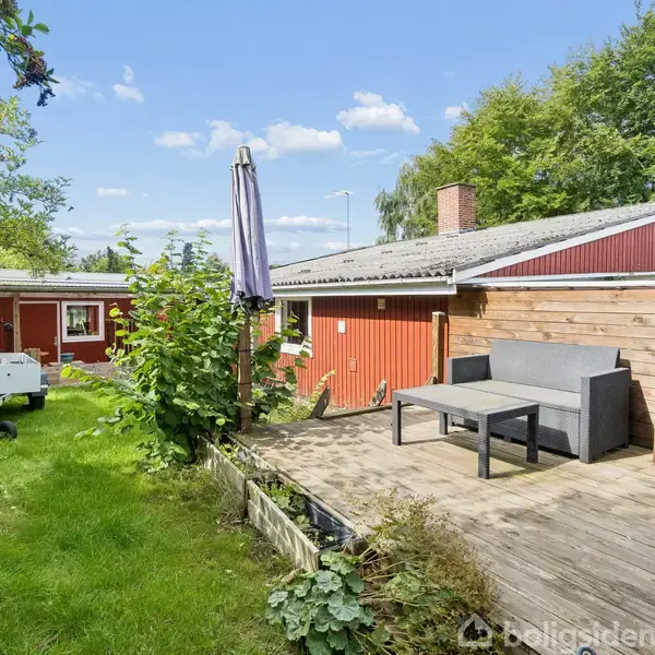 Terrasse med grå sofa og parasol står ved siden af en rød træbygning. Græsplæne og buske omkranser området, og en trailer er placeret til venstre.
