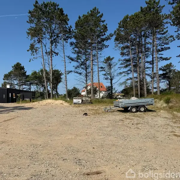 En trailer står stille på en grusplads omgivet af høje træer. I baggrunden ses en hvid bygning med rødt tag samt en sort bygning.