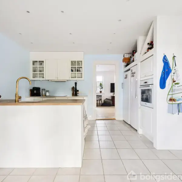 Et moderne køkken med hvidt interiør, en central ø med en gylden vandhane og blender, samt en indbygget ovn og mikrobølgeovn. Der hænger tre viskestykker på væggen.