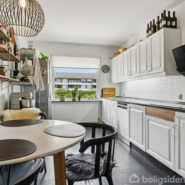 Et lyst køkken med hvide skabe og sorte bordplader. Et rundt bord med to stole står centralt placeret. Ud af vinduet ses en boligblok. En moderne lampe hænger fra loftet.