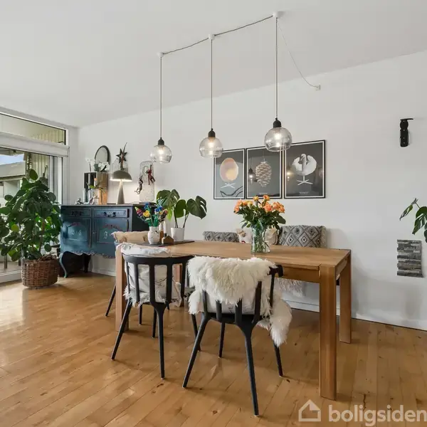 Et spisebord med stole er centreret i et lyst rum med trægulv. Loftslamper hænger over bordet, og grønne planter står rundt omkring. Store vinduer åbner ud til en terrasse.