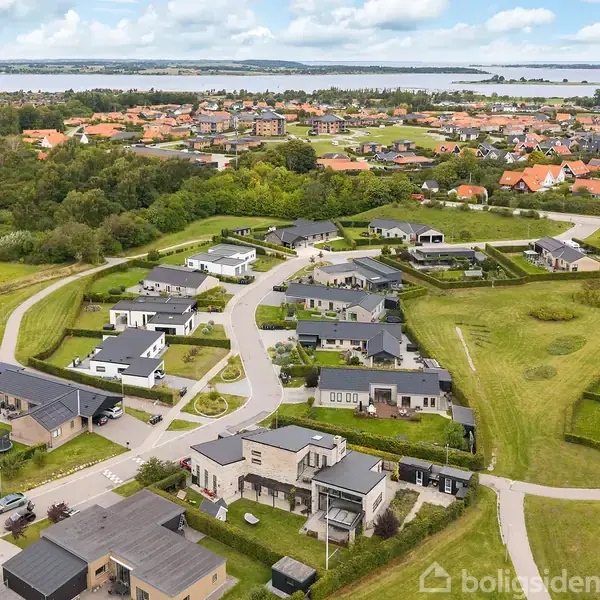 Flere moderne huse står langs snoede veje i en grøn forstad med græsplæner og træer. I baggrunden ses en større by samt en sø eller fjord.