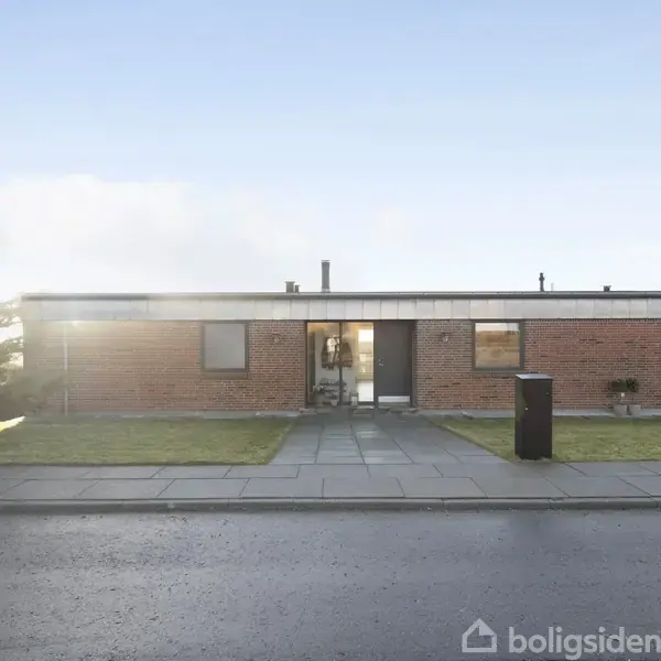 En lav, rektangulær bygning lavet af mursten med fladt tag står stille ved en gade. Træer og græsplæne omgiver huset i en rolig forstadsmiljø.