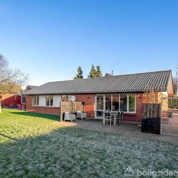 Et rødt murstenshus med en grå tag, der står på en solrig, frodig græsplæne. Havemøbler er placeret foran en terrasse, omkranset af træer i en villahave.