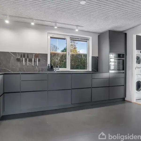Moderne køkken med mørke skabe, malede vægge, og en sort marmor bordplade. I venstre hjørne står en blomstervase. Gennem vinduet ses en have. Der er også en vaskerum i baggrunden.