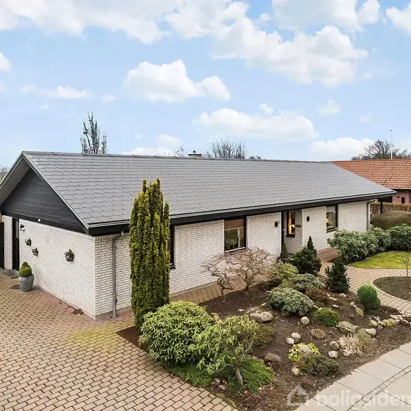 Et hvidt parcelhus med sort tag og garage ved siden af, omgivet af en velplejet have. Flisebelagt indkørsel leder op til hoveddøren. Baggrund viser åbne marker.