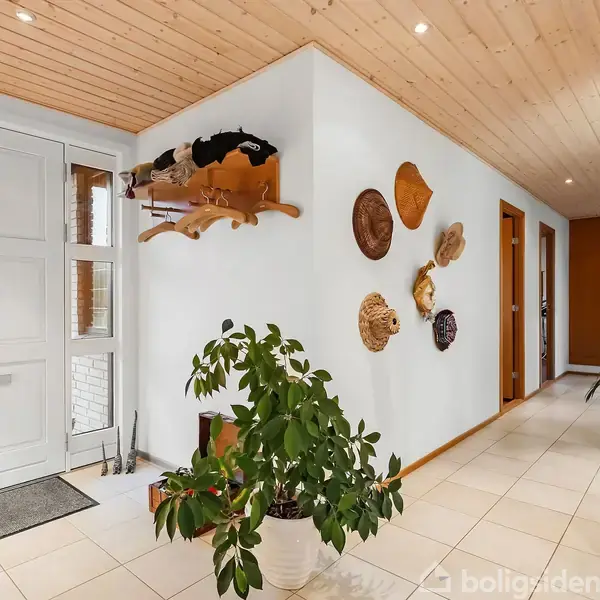 Dør med glasruder åbner til en lys gang med hvidmalede vægge og flisegulv. Planter i potter og hatte hænger på væggen. Træloft med indbyggede spotlights.