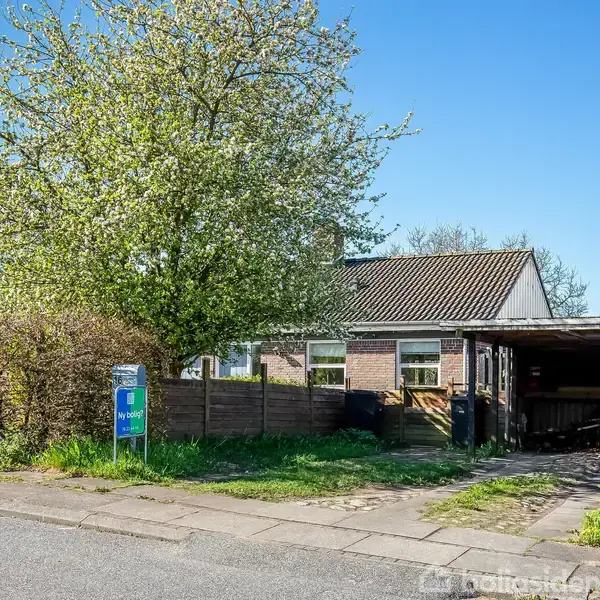 Et hus med en carport, omkranset af grønne buske og et blomstrende træ. Et skilt ved vejen viser teksten "Til salg" og "Nybolig."