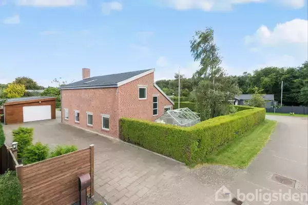 Et rødt murstenshus med en skrå tag og flere vinduer står stille. Huset er omgivet af en velplejet hæk og en indkørsel, mens en garage er i baggrunden.