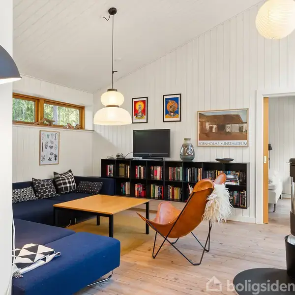 En stue med blå sofa og læderstol omkring et træbord. Reol med bøger, vægkunst, fladskærm og en døråbning til et andet rum. Lyse vægge og trægulv.