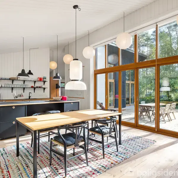 Køkkenområde med træbord og sorte stole, hvor lamper hænger over. Store vinduer viser en terrasse med udemøbler og skov. Et farverigt tæppe ligger på gulvet.