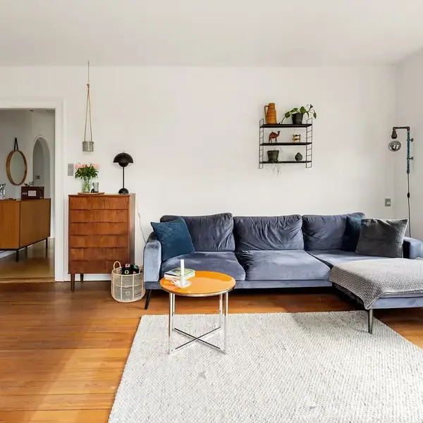 Blå sofa står i stue ved gråt tæppe og lille træbord. Væggene er hvide med en hylde over sofaen. Et vindue og planter omgiver rummet, som er lyst og moderne.