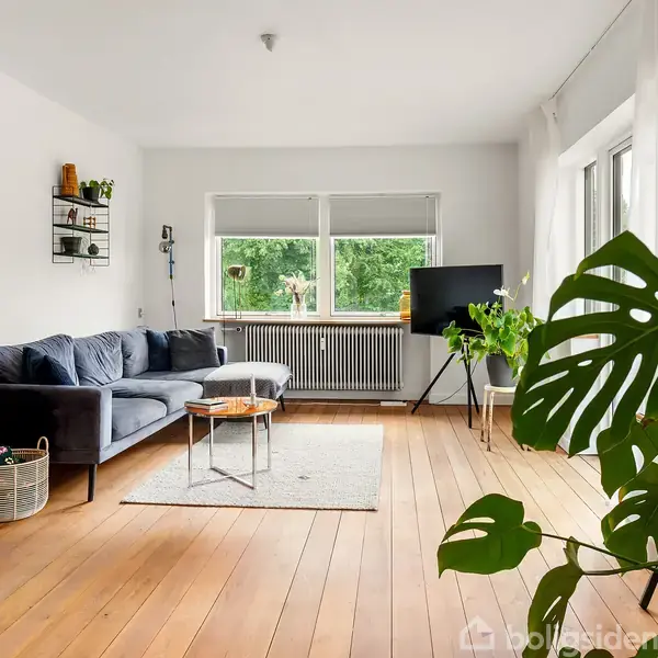 En moderne stue indeholder en grå sofa ved siden af en trækommode. Et sofabord står på et tæppe, omgivet af grønne planter. Store vinduer giver udsigt til en grøn have.