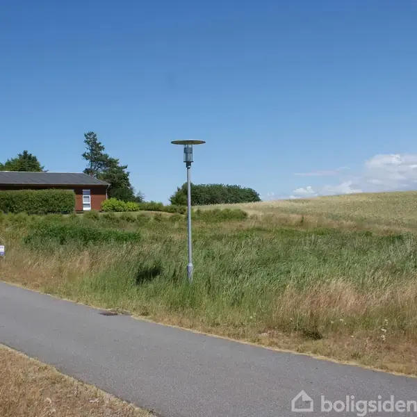 Et rødt hus uden bevægelse er beliggende på en bakketop ved siden af en asfaltvej. Der er et skilt foran huset. Omgivet af højt græs og træer.